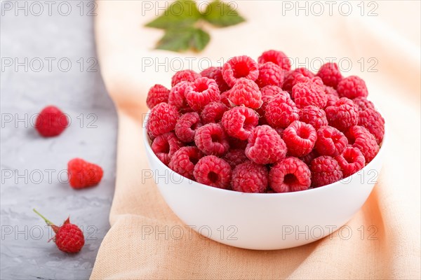 Fresh raspberry in white bowl and orange pastel textile on gray concrete background. side view, close up, selective focus