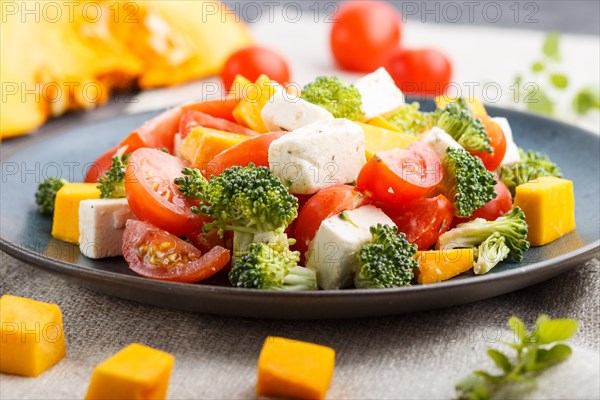 Vegetarian salad with broccoli, tomatoes, feta cheese, and pumpkin on a blue ceramic plate on a black concrete background and linen textile, side view, close up, selective focus