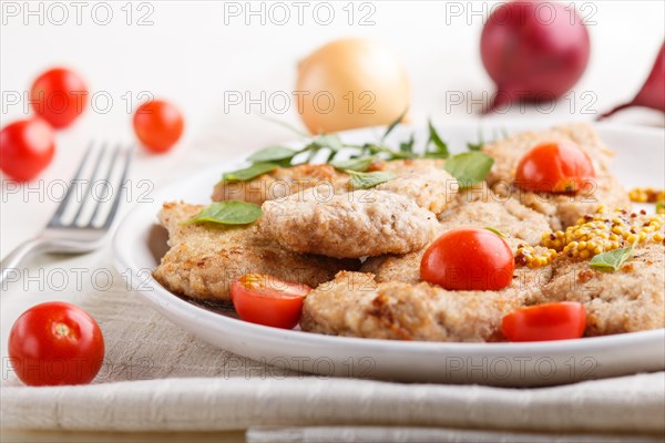 Fried pork chops with tomatoes and herbs on a white ceramic plate on a white wooden background and linen textile. side view, close up, selective focus