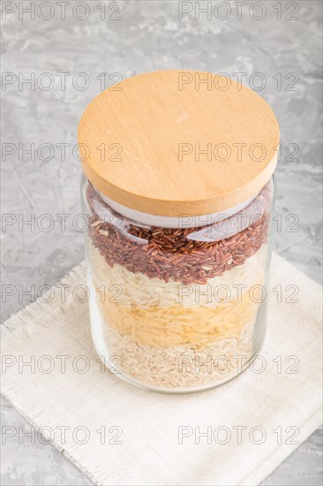 Glass jar with different kinds of rice poured in layers on a gray concrete background. side view, close up. Healthy food concept