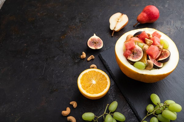 Vegetarian fruit salad of watermelon, grapes, figs, pear, orange, cashew on slate board on a black concrete background. Side view, copy space