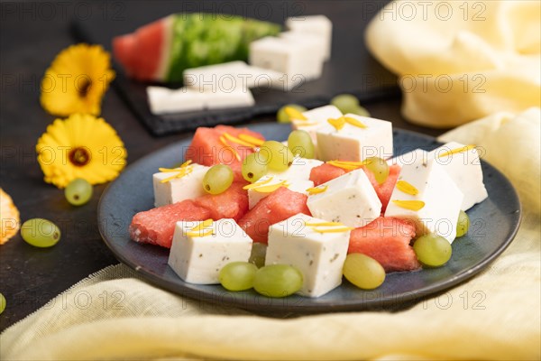 Vegetarian salad with watermelon, feta cheese, and grapes on blue ceramic plate on black concrete background and yellow linen textile. Side view, close up, selective focus