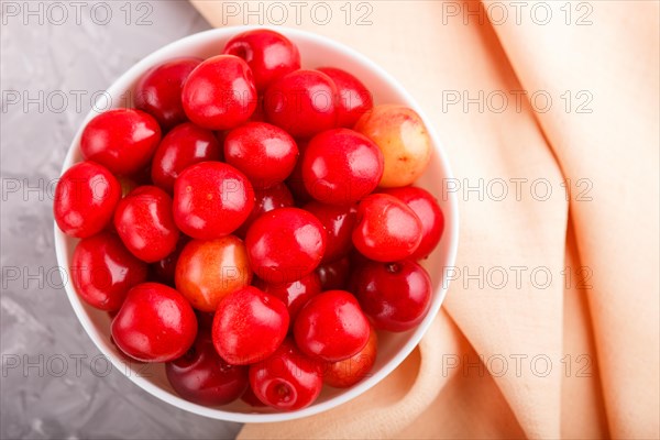 Fresh red sweet cherry in white bowl on gray background with orange textile. top view, flat lay