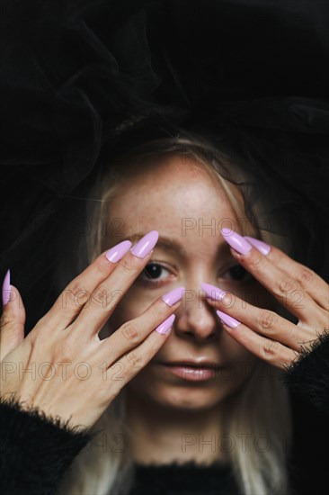 A striking image of a young blonde caucasian woman with purple nails covering her face with both hands, wear a vintage black lace dress and hat, looking at camera