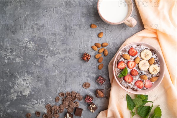 Chocolate cornflakes with milk, strawberry and almonds in ceramic bowl on gray concrete background and orange linen textile. Top view, flat lay, copy space