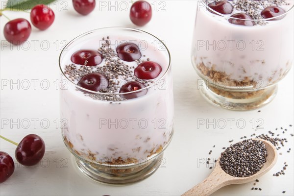 Yoghurt with cherries, chia seeds and granola in glass with wooden spoon on white wooden background. side view, close up
