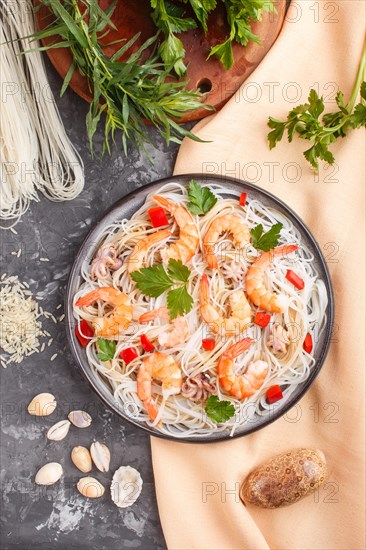 Rice noodles with shrimps or prawns and small octopuses on gray ceramic plate on a black concrete background and orange textile. Top view, flat lay, close up