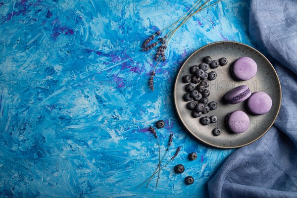Purple macarons or macaroons cakes with blueberries on ceramic plate on a blue concrete background and blue textile. Flat lay, top view, copy space