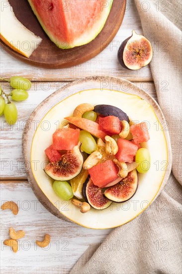 Vegetarian fruit salad of watermelon, grapes, figs, pear, orange, cashew on white wooden background and linen textile. Top view, flat lay, close up
