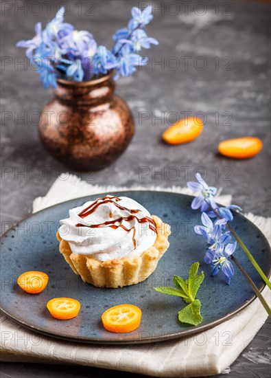 Cake with whipped egg cream on a blue ceramic plate with kumquat slices and mint leaves on a black concrete background with linen napkin. decorated with blue flowers. side view
