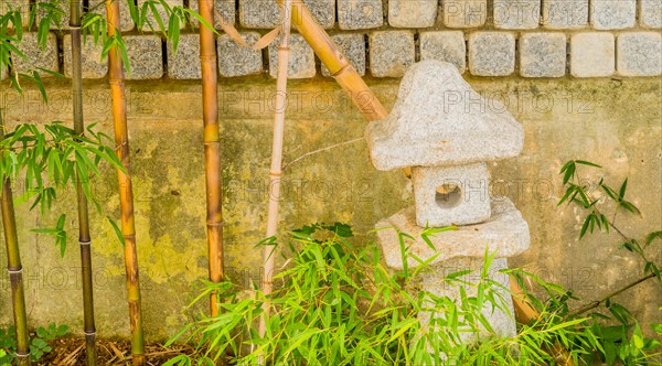 Small cement spirit house in front of concrete wall next to young bamboo plants in South Korea