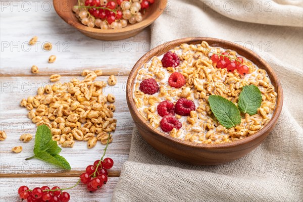 Wheat flakes porridge with milk, raspberry and currant in wooden bowl on white wooden background and linen textile. Side view, close up