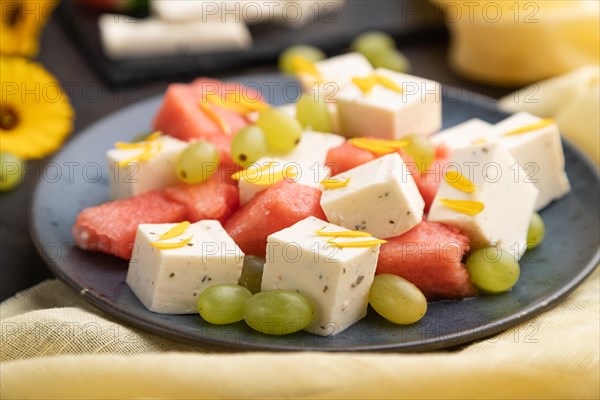 Vegetarian salad with watermelon, feta cheese, and grapes on blue ceramic plate on black concrete background and yellow linen textile. Side view, close up, selective focus