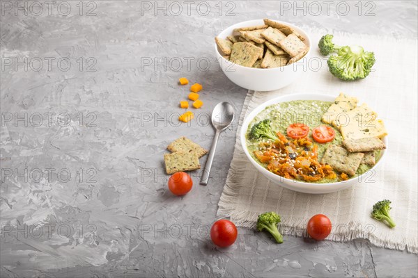 Green broccoli cream soup with crackers and cheese in white bowl on a gray concrete background and linen napkin. side view, copy space