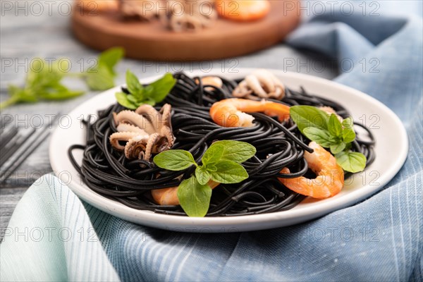 Black cuttlefish ink pasta with shrimps or prawns and small octopuses on gray wooden background and blue textile. Side view, close up, selective focus