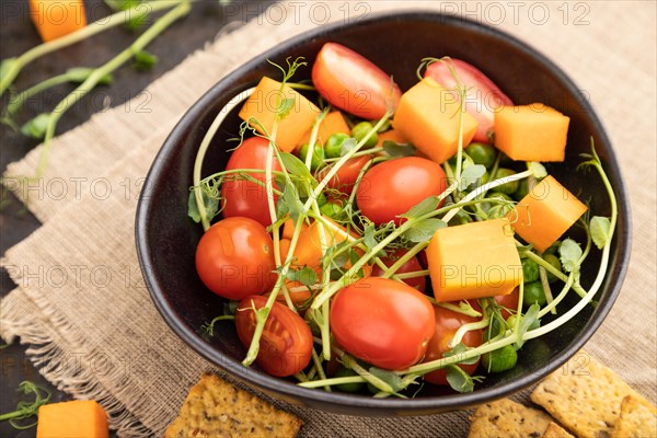 Vegetarian vegetable salad of tomatoes, pumpkin, microgreen pea sprouts on black concrete background and linen textile. Side view, close up, selective focus