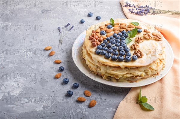 Homemade layered Napoleon cake with milk cream. Decorated with blueberry, almonds, walnuts, hazelnuts, mint on a gray concrete background and orange textile. side view. copy space