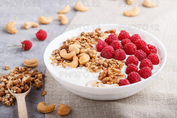 Yoghurt with raspberry, granola, cashew and walnut in white plate with wooden spoon on gray concrete background and linen textile. side view, close up, selective focus