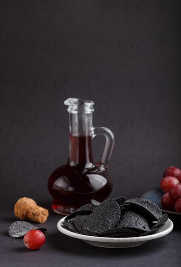 Black potato chips with charcoal, balsamic vinegar in glass, red grapes on a blue ceramic plate on a black background. side view, close up, selective focus, copy space