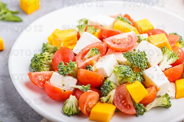 Vegetarian salad with broccoli, tomatoes, feta cheese, and pumpkin on white ceramic plate on a gray concrete background and linen textile, side view, close up, selective focus