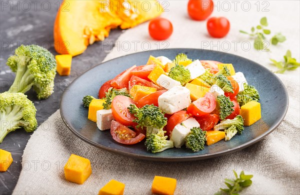 Vegetarian salad with broccoli, tomatoes, feta cheese, and pumpkin on a blue ceramic plate on a black concrete background and linen textile, side view, close up, selective focus