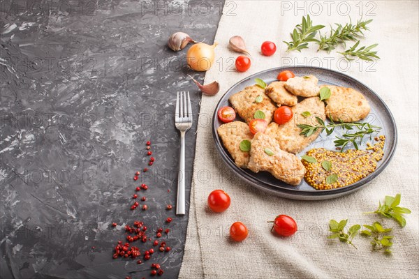 Fried pork chops with tomatoes and herbs on a gray ceramic plate on a black concrete background and linen textile. side view, copy space