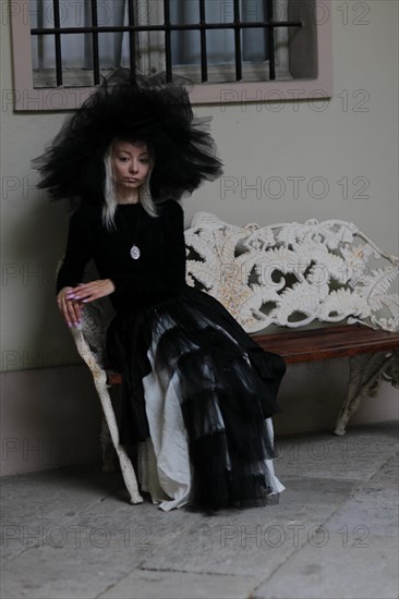 Gothically styled white thin blonde woman sitting on a lace-covered bench in a vintage indoor setting, wear lace veil and hat, black and white dress