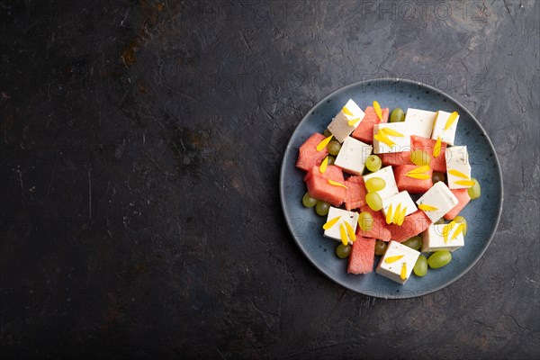 Vegetarian salad with watermelon, feta cheese, and grapes on blue ceramic plate on black concrete background. top view, copy space, flat lay