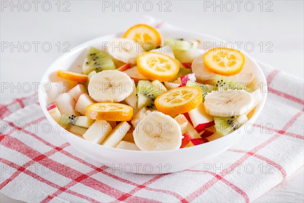 Vegetarian salad of bananas, apples, pears, kumquats and kiwi on linen tablecloth, close up, selective focus. white wooden background