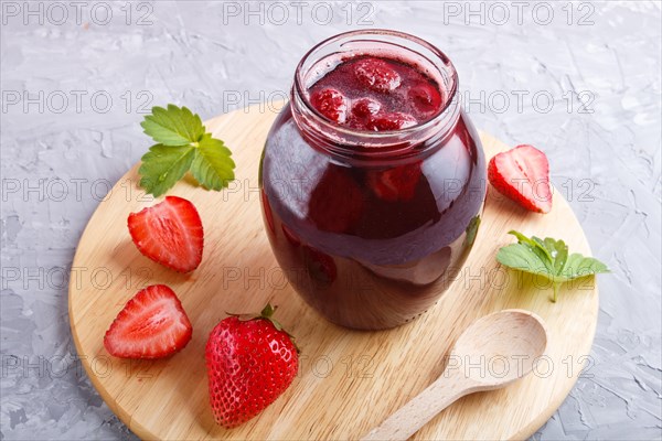 Strawberry jam in a glass jar with berries and leaves on gray concrete background. Homemade, close up, side view, selective focus