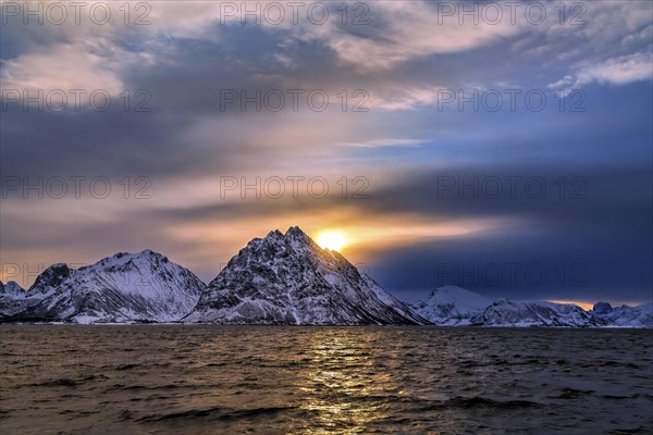 Blick auf die verschneiten Berge mit tiefstehender Sonne und einem tollen Wolkenhimmel auf den Vesteralen in Norwegen und dem Nordmeer im Vordergrund