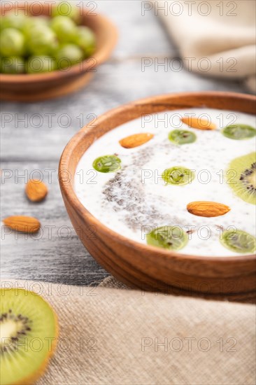 Yogurt with kiwi, gooseberry, chia and almonds in wooden bowl on gray wooden background and linen textile. Side view, close up, selective focus