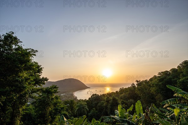 View from a mountain onto a secluded bay with a sandy beach and mangrove forest. The sun sets over the sea and boats float in the water. Grande Anse beach, Basse Terre, Guadeloupe, French Antilles, Caribbean, North America