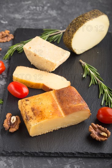Smoked cheese and various types of cheese with rosemary and tomatoes on black slate board on a black concrete background. Side view, close up, selective focus