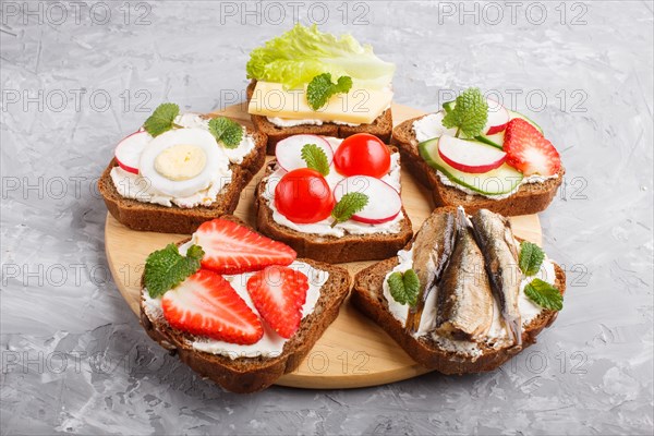 Set of different sandwiches with cheese, radish, lettuce, strawberry, sprats, tomatoes and cucumber on wooden board on a gray concrete background. side view, close up