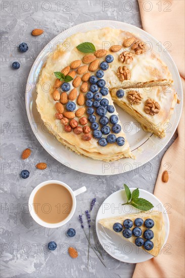 Homemade layered Napoleon cake with milk cream. Decorated with blueberry, almonds, walnuts, hazelnuts, mint on a gray concrete background and cup of coffee. top view. flat lay