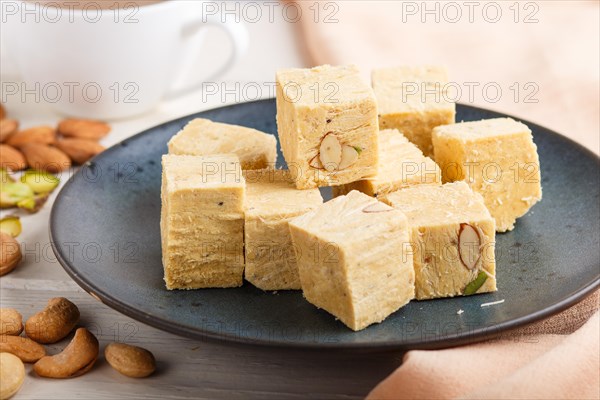 Traditional indian candy soan papdi in a blue ceramic plate with almond, pistache, cashew and a cup of coffee on a white wooden background with orange textile. side view, close up, selective focus