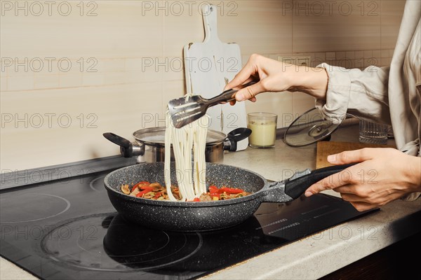 Unrecognizable woman transfers noodles with kitchen tongs into a frying pan with vegetable dressing and beef