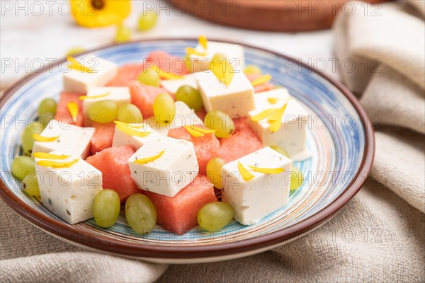 Vegetarian salad with watermelon, feta cheese, and grapes on blue ceramic plate on white wooden background and linen textile. Side view, close up, selective focus