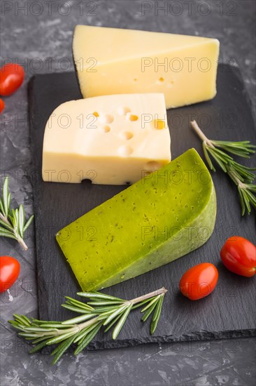 Green basil cheese and various types of cheese with rosemary and tomatoes on black slate board on a black concrete background. Side view, close up, selective focus