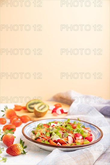 Vegetarian fruits and vegetables salad of strawberry, kiwi, tomatoes, microgreen sprouts on white and orange background and linen textile. Side view, copy space, selective focus