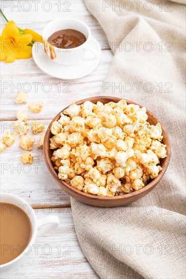 Popcorn with caramel in wooden bowl and a cup of coffee on a white wooden background and linen textile. Side view, close up