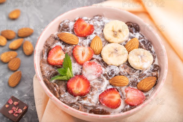 Chocolate cornflakes with milk, strawberry and almonds in ceramic bowl on gray concrete background and orange linen textile. Side view, close up, selective focus