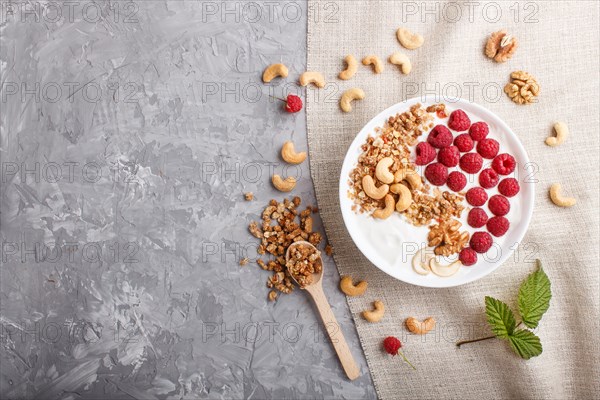 Yoghurt with raspberry, granola, cashew and walnut in white plate with wooden spoon on gray concrete background and linen textile. top view, flat lay, copy space