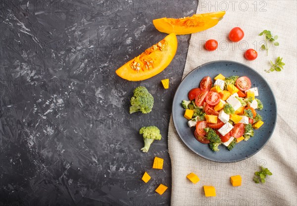 Vegetarian salad with broccoli, tomatoes, feta cheese, and pumpkin on a blue ceramic plate on a black concrete background and linen textile, top view, copy space, flat lay