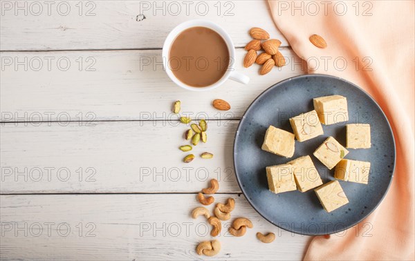 Traditional indian candy soan papdi in a blue ceramic plate with almond, pistache, cashew and a cup of coffee on a white wooden background with orange textile. top view, flat lay, copy space