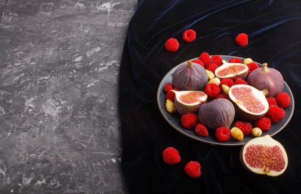 Fresh figs, strawberries and raspberries on blue ceramic plate on black concrete background and blue velvet textile. side view, copy space