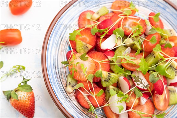 Vegetarian fruits and vegetables salad of strawberry, kiwi, tomatoes, microgreen sprouts on white concrete background and linen textile. Top view, close up, selective focus