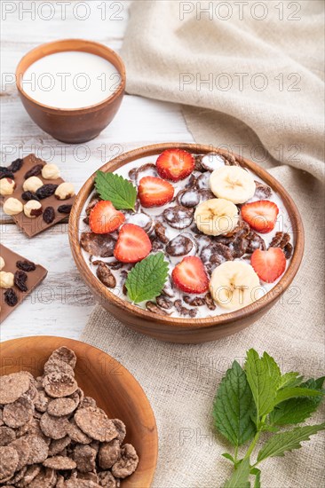 Chocolate cornflakes with milk and strawberry in wooden bowl on white wooden background and linen textile. Side view, close up