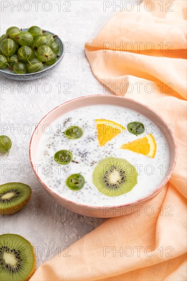 Yogurt with kiwi, gooseberry, chia in ceramic bowl on gray concrete background and orange linen textile. Side view, close up